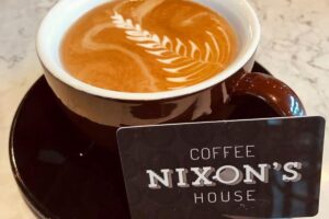 A galss coffee mug with coffee inside next to a Nixon's House sign